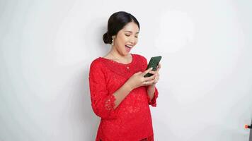 en leende asiatisk kvinna bär röd kebaya och pannband och innehav henne telefon, isolerat förbi vit bakgrund. Indonesiens oberoende dag begrepp video