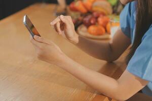 A young woman with a beautiful face in a blue shirt with long hair eating fruit sitting inside the kitchen at home with a laptop and notebook for relaxation, Concept Vacation. photo