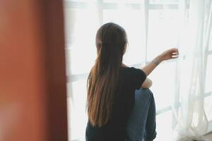 Thoughtful girl sitting on sill embracing knees looking at window, sad depressed teenager spending time alone at home, young upset pensive woman feeling lonely or frustrated thinking about problems photo