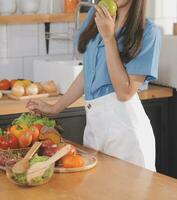 A young woman with a beautiful face in a blue shirt with long hair eating fruit sitting inside the kitchen at home with a laptop and notebook for relaxation, Concept Vacation. photo