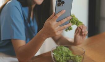 A young woman with a beautiful face in a blue shirt with long hair eating fruit sitting inside the kitchen at home with a laptop and notebook for relaxation, Concept Vacation. photo