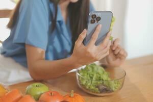 A young woman with a beautiful face in a blue shirt with long hair eating fruit sitting inside the kitchen at home with a laptop and notebook for relaxation, Concept Vacation. photo