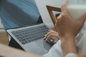 Happy young female in casual clothes smiling and looking at camera while using laptop on bed in bedroom at home photo