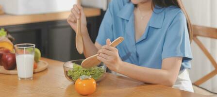 A young woman with a beautiful face in a blue shirt with long hair eating fruit sitting inside the kitchen at home with a laptop and notebook for relaxation, Concept Vacation. photo