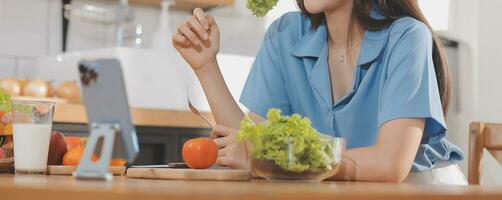 A young woman with a beautiful face in a blue shirt with long hair eating fruit sitting inside the kitchen at home with a laptop and notebook for relaxation, Concept Vacation. photo