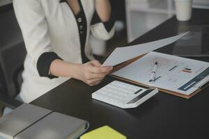 mujer de negocios asiática que usa una computadora portátil y trabaja en la oficina con un documento de calculadora en el escritorio, planificando el análisis del informe financiero, la inversión del plan de negocios, el concepto de análisis financiero. foto