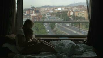 View of mother lying on the bed with small son against huge panoramic window and cityscape, Barcelona, Spain video