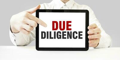Text DUE DILIGENCE on tablet display in businessman hands on the white background. Business concept photo