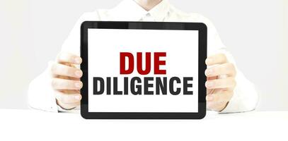 Text DUE DILIGENCE on tablet display in businessman hands on the white background. Business concept photo