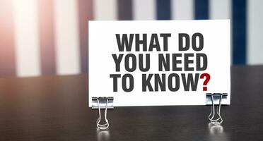 WHAT DO YOU NEED TO KNOW sign on paper on dark desk in sunlight. Blue and white background photo