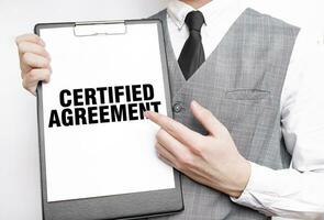 CERTIFIED AGREEMENT inscription on a notebook in the hands of a businessman on a gray background, a man points with a finger to the text photo