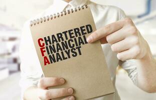 Text CHARTERED FINANCIAL ANALYST on brown paper notepad in businessman hands in office. Business concept photo