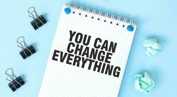 White notepad with text YOU CAN CHANGE EVERYTHING and office tools on the blue background photo
