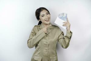 A smiling young government worker woman is wearing khaki uniform holding cash money in Indonesian rupiah isolated by white background photo