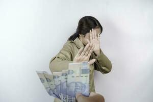 A serious young government worker woman is wearing khaki uniform gesturing rejection to cash money in Indonesian rupiah isolated by white background photo