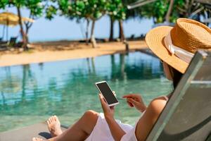 Young woman traveler relaxing and using a mobile phone by a resort pool while traveling for summer vacation, Travel lifestyle concept photo