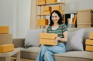 puesta en marcha pequeño negocio pequeño, empresario propietario mujer utilizando teléfono inteligente o tableta tomando recibir y comprobación en línea compra compras orden a preparando paquete producto caja. en sofá foto