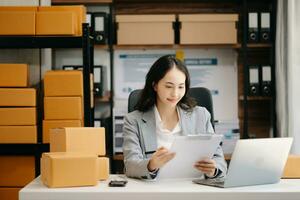 Startup small business SME, Entrepreneur owner woman using smartphone or tablet taking receive and checking online purchase shopping order to preparing pack product box. photo