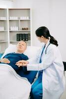 asiático médico en blanco traje tomar notas mientras que se discute y asiático anciano, hombre paciente quien acostado en cama con recepción salina solución en hospital foto