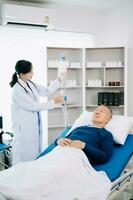 asiático médico en blanco traje tomar notas mientras que se discute y asiático anciano, hombre paciente quien acostado en cama con recepción salina solución en hospital foto
