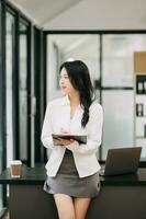 confidente negocio experto atractivo sonriente joven asiático mujer participación digital tableta y ordenador portátil en escritorio en oficina. foto