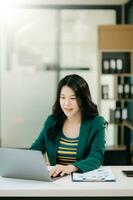 Beautiful Asian woman using laptop and tablet while sitting at her working place. Concentrated at work. in office photo