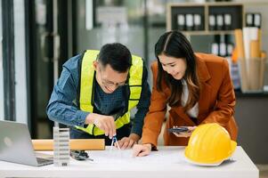 Contractor and architect looking at plan, tablet, laptop and talking about working on big project. Building in construction office interior. photo