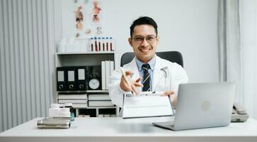 médico médico sentar en mesa en hospital. atractivo terapeuta masculino facultativo farmacéutico sonrisa y utilizar tableta y ordenador portátil. después éxito trabajo desde tratamiento foto