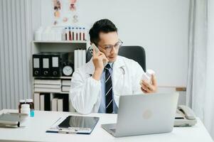 Asian Doctor talking over his mobile phone and making notes in a note. Medicine professional talking on phone and clinical report test results photo