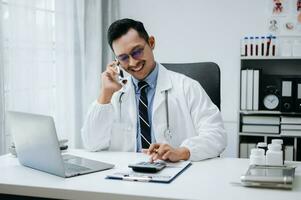 confidente joven asiático masculino médico en blanco médico uniforme sentar a escritorio trabajando en computadora. sonriente utilizar ordenador portátil escribir en médico diario en clínica. foto