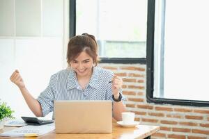 hermosa joven adolescente asiático negocio mujer utilizando computadora ordenador portátil con manos arriba en ganador es un gesto, contento a ser exitosamente celebrando logro éxito. foto