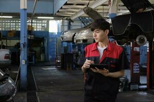 One male Asian professional mechanic, supervisor engineer, inspects repair work checklists with tablet at garage, service car maintenance, and fixing specialist occupations in auto transport industry. photo