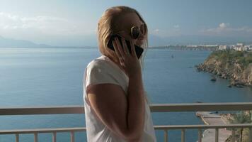 een blond haar vrouw pratend naar een telefoon Aan een hotel balkon in de buurt de zee landschap video