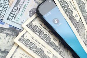 Smartphone screen with Tiktok app and lot of hundred dollar bills. Business and social networking concept photo