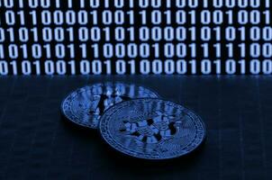 A pair of bitcoins lies on a cardboard surface on the background of a monitor depicting a binary code of bright zeros and one units on a black background. phantom classic blue color. photo