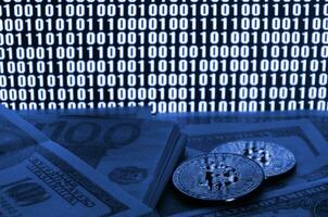 Two bitcoins lies on a pile of dollar bills on the background of a monitor depicting a binary code of bright zeros and one units on a black background. phantom classic blue color photo