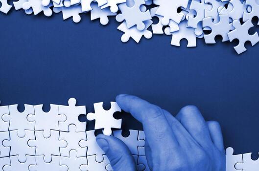 The hand folds a white jigsaw puzzle and a pile of uncombed puzzle pieces lies against the background of phantom classic blue color surface. Texture photo with space for text