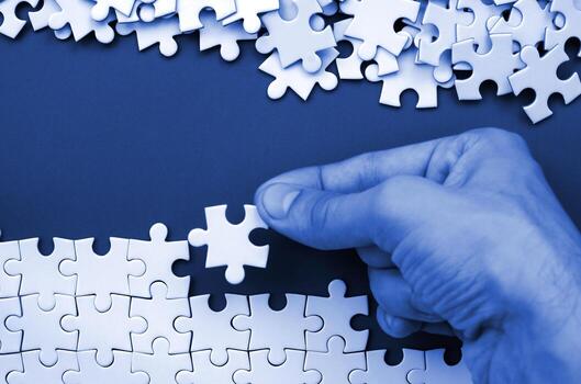 The hand folds a white jigsaw puzzle and a pile of uncombed puzzle pieces lies against the background of phantom classic blue color surface. Texture photo with space for text