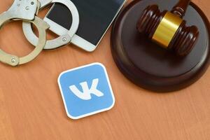 Vkontakte paper logo lies with wooden judge gavel, smartphone and handcuffs. Entertainment lawsuit concept photo