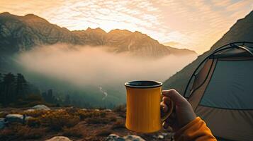 Camping holding a cup in an open yellow tent with a misty mountain lit by natural light. photo