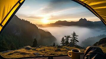 Camping holding a cup in an open yellow tent with a misty mountain lit by natural light. photo