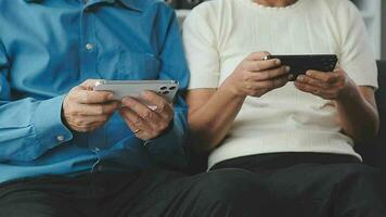 Happy senior old couple holding smartphone looking at cellphone screen laughing relaxing sit on sofa together, smiling elder mature grandparents family embracing having fun with mobile phone at home video