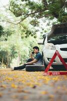 Asian man sitting beside car and using mobile phone calling for assistance after a car breakdown on street. Concept of vehicle engine problem or accident and emergency help from Professional mechanic photo