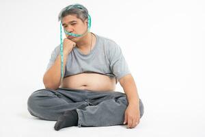 An overweight man sitting on the floor and Bored after uses a measurement tape for measures his fat belly. isolated on white background. Concept of Healthcare, medicine, and  Weight loss photo