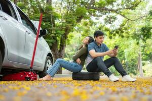 Asian couple using smartphone for assistance after a car breakdown on street. Concept of vehicle engine problem or accident and emergency help from Professional mechanic photo
