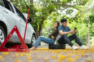 asiático Pareja utilizando teléfono inteligente para asistencia después un coche Descompostura en calle. concepto de vehículo motor problema o accidente y emergencia ayuda desde profesional mecánico foto