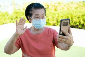 Happy Asian elderly wearing a face mask and answering a Video Call Using her Smartphone in the park and chatting with family, Technology and older people concept. photo