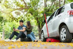 asiático Pareja utilizando teléfono inteligente para asistencia después un coche Descompostura en calle. concepto de vehículo motor problema o accidente y emergencia ayuda desde profesional mecánico foto