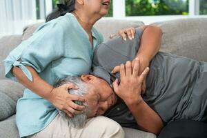 Asian old elderly man is In pain with his hands on his chest and having a heart attack in the Living room and his wife is shocked and confused, helpless. Concept of healthcare and Elderly caregivers photo