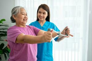 Happy elderly female exercising with rubber expander with Young physiotherapist for recovery arm muscles in a physiotherapy center. Concept of happy retirement with care from a caregiver and health. photo